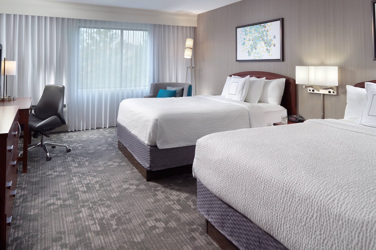 A hotel room with two beds, a desk, a chair, and a large window with sheer curtains.