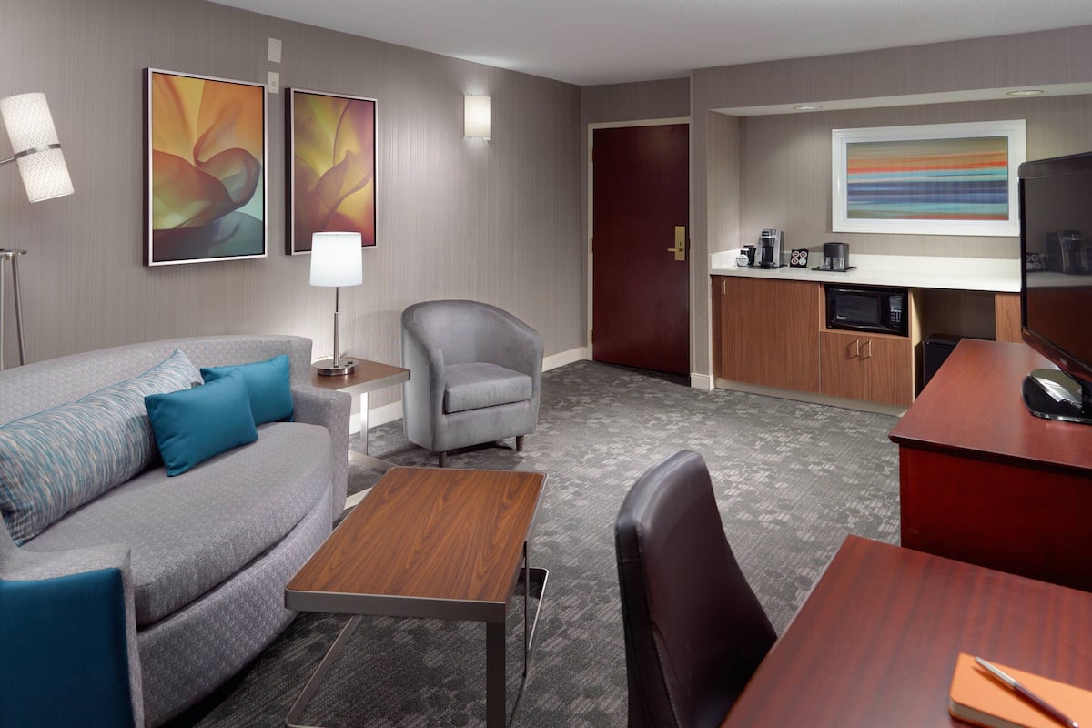 A hotel room with a grey sofa, a wooden table, a grey armchair, a desk, a microwave, a coffee maker, and a TV.