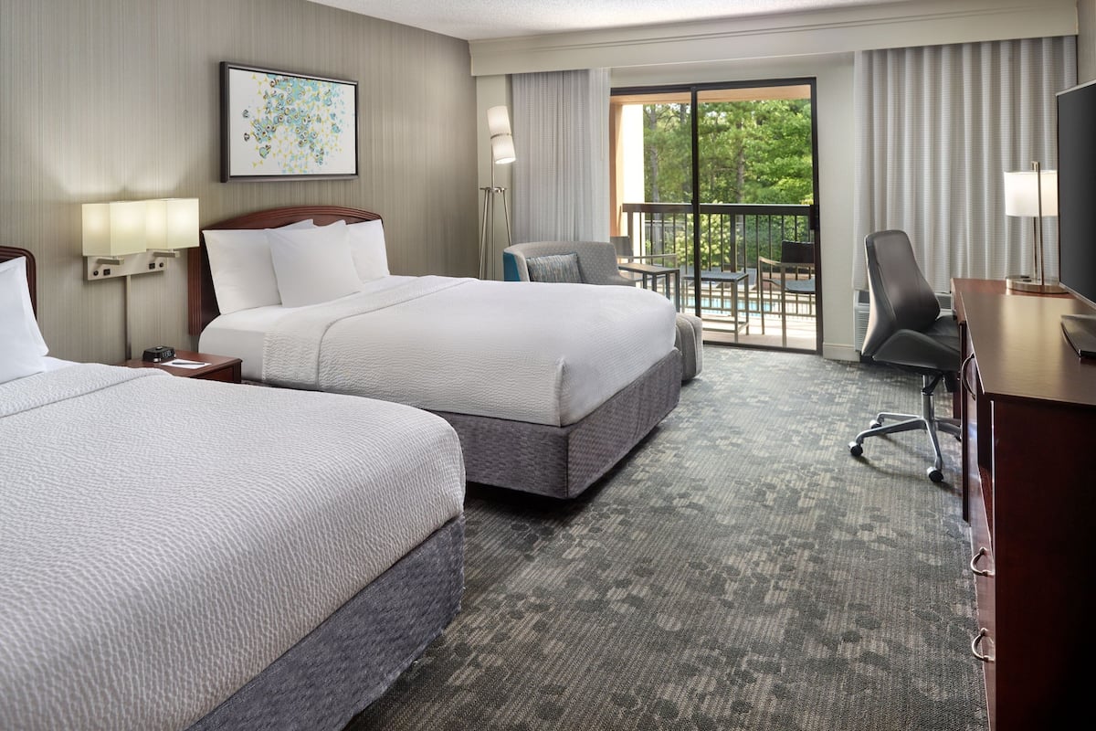 A hotel room with two beds, a desk, a chair, and a balcony with a view of trees.
