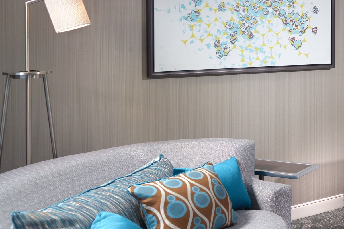 A modern living room with a grey sofa, blue and patterned cushions, a floor lamp, and a framed abstract painting on the wall.