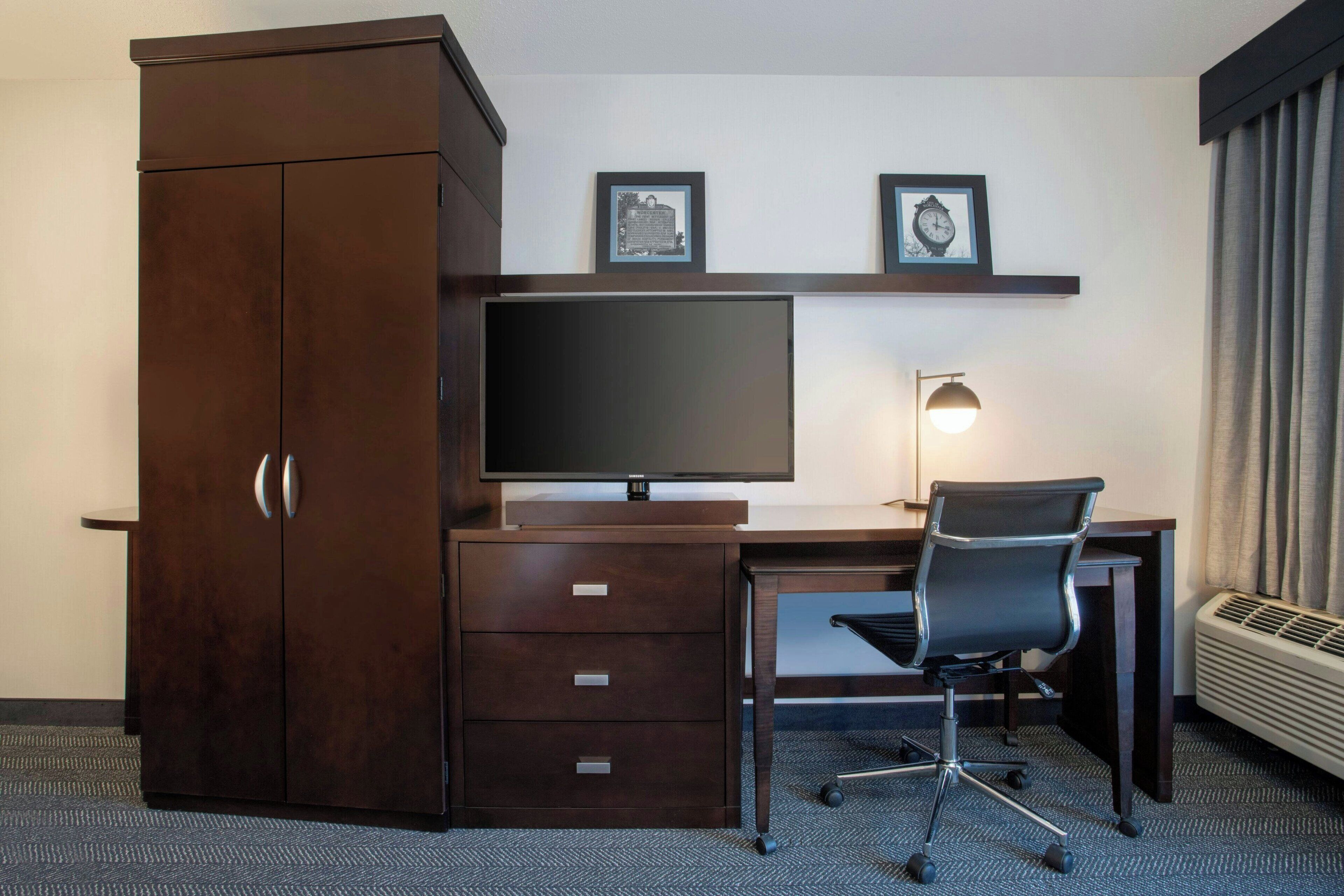 A hotel room with a dark wooden desk, a flat-screen TV, a black office chair, a desk lamp, and a wardrobe.