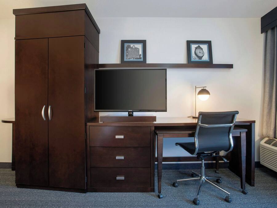 A hotel room with a dark wooden desk, a flat-screen TV, a black office chair, a desk lamp, and a wardrobe.