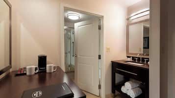 Habitación de hotel con un escritorio de madera, una computadora portátil, dos tazas, una cafetera, un lavamanos, un espejo y una puerta que da a la zona de la ducha.