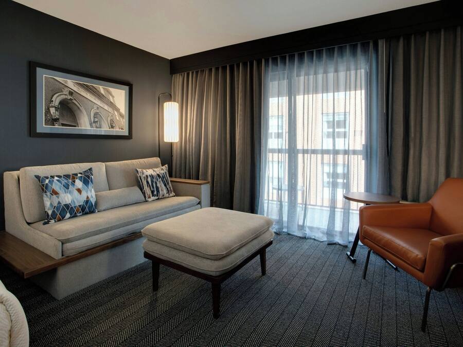 A modern hotel room with a sofa, armchair, ottoman, and a framed picture on the wall.