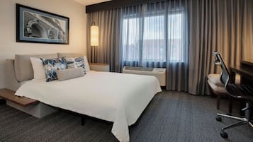 A hotel room with a bed, a desk, an air conditioning unit, a chair, and a large window with curtains.