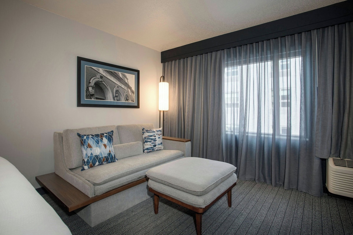 A hotel room with a sofa, ottoman, and a framed picture on the wall.