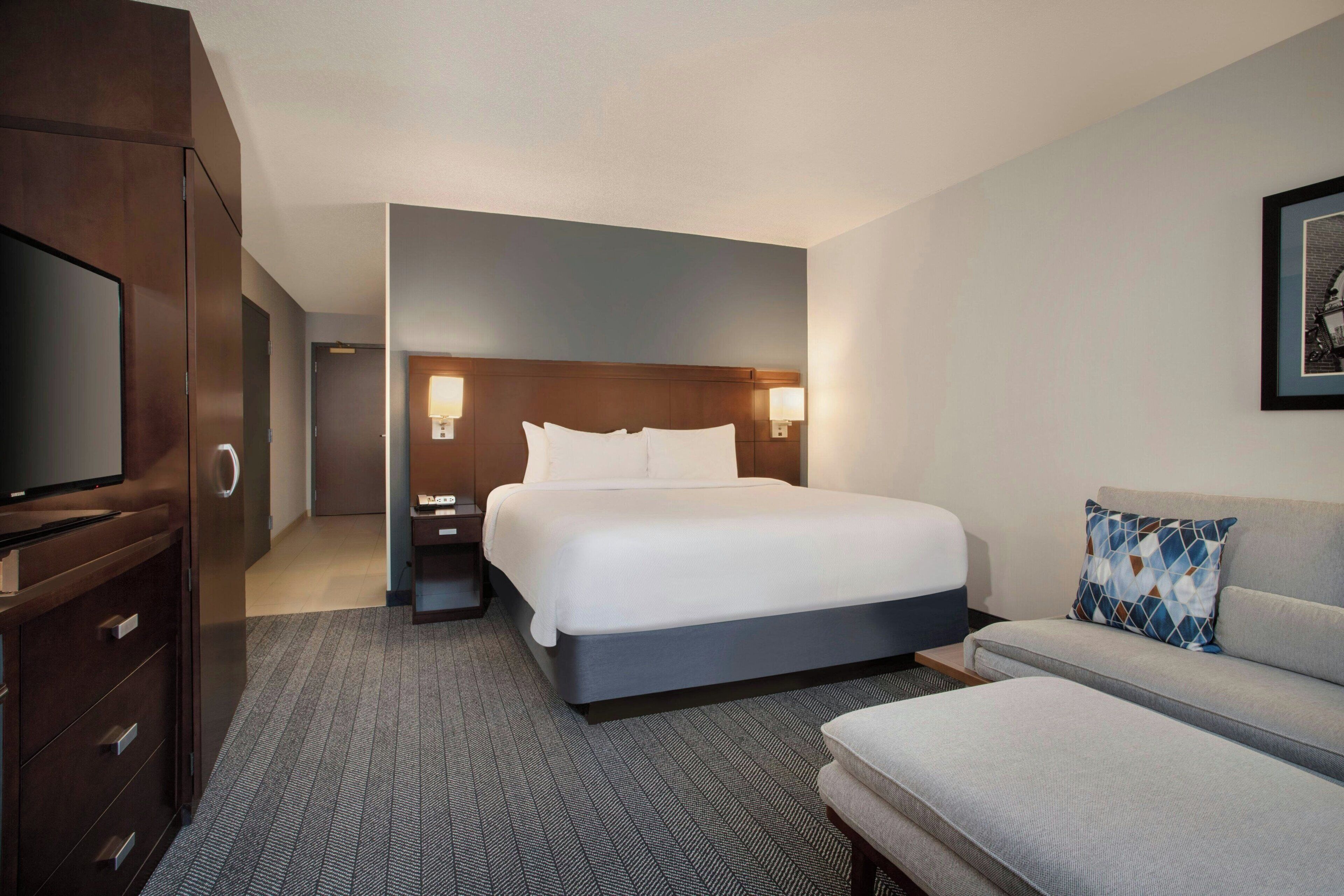 Quarto de hotel com uma cama grande, televisão, sofá e mesa de cabeceira com luminárias.