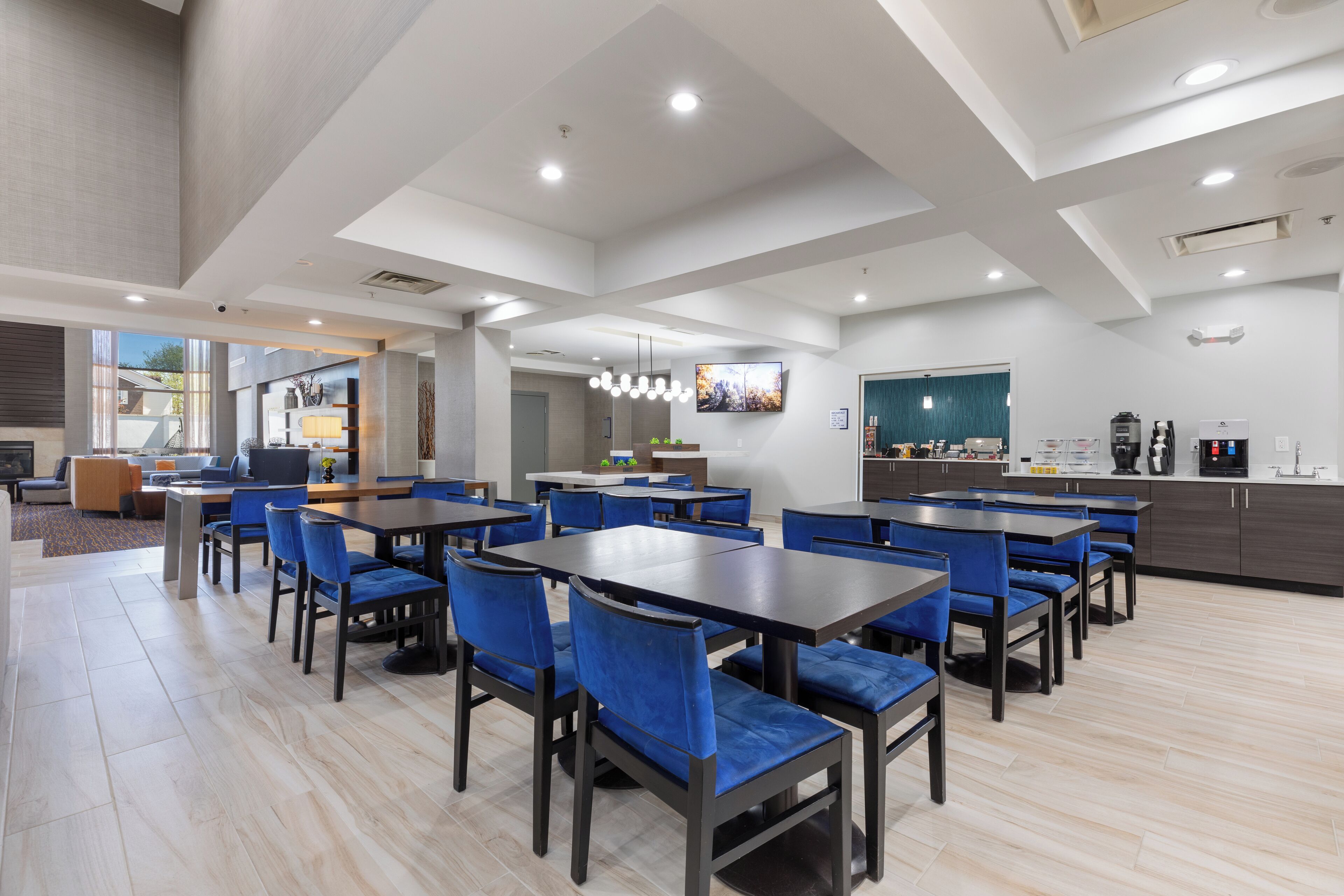 Un espace repas moderne avec plusieurs tables et des chaises bleues, un coin cuisine équipé d’une machine à café, et un grand écran de télévision fixé au mur.