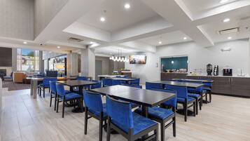 Un espace repas moderne avec plusieurs tables et des chaises bleues, un coin cuisine équipé d’une machine à café, et un grand écran de télévision fixé au mur.