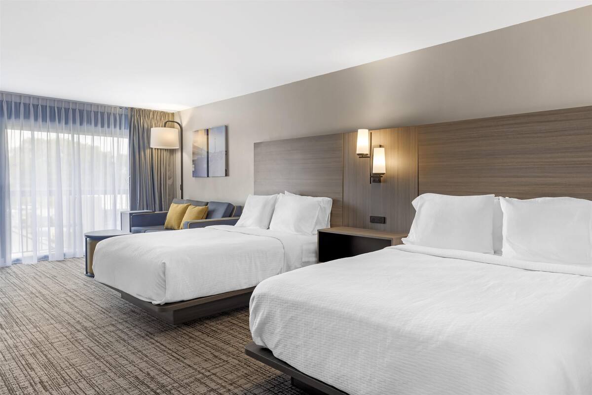 Habitación de hotel con dos camas, un ventanal con cortinas traslúcidas, una lámpara de pared y un cuadro.