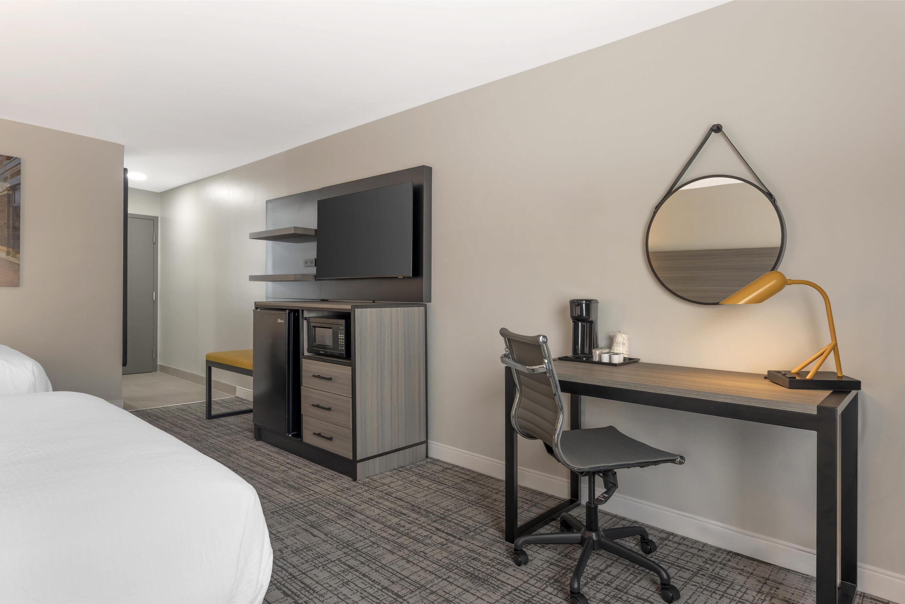A hotel room with a bed, a desk with a chair, a television, a coffee maker, and a mirror on the wall.