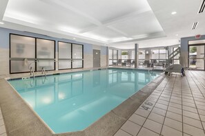 Una amplia piscina interior con agua cristalina, paredes enlosadas y una zona de estar con mesas y sillas.