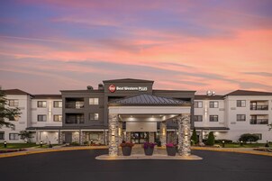 Un hotel Best Western PLUS con diseño moderno, que cuenta con una entrada central con galería techada, pilares de piedra y plantas en macetas.