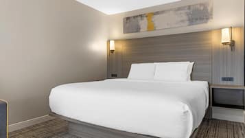 Quarto de hotel moderno com uma cama grande, cabeceira com iluminação integrada, obra de arte na parede e mesa de cabeceira com abajur.