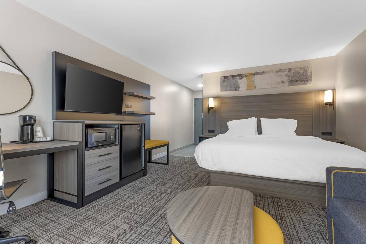 Una habitación de hotel moderna con una cama, un televisor de pantalla plana, un microondas, una cafetera, un escritorio y una mesita pequeña.