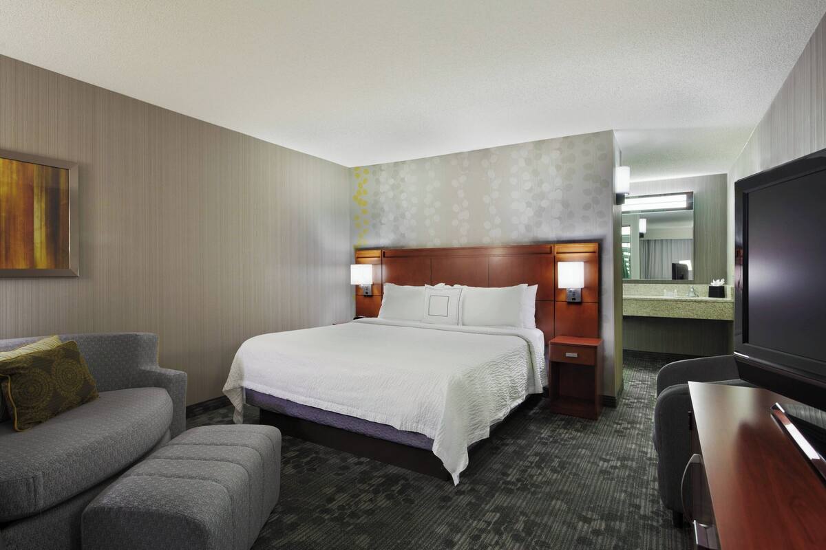Quarto de hotel com cama, sofá, televisão, escrivaninha e banheiro com espelho.