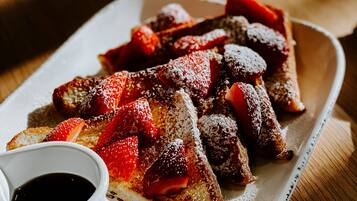 A plate of French toast with strawberries and powdered sugar, accompanied by a small cup of syrup.