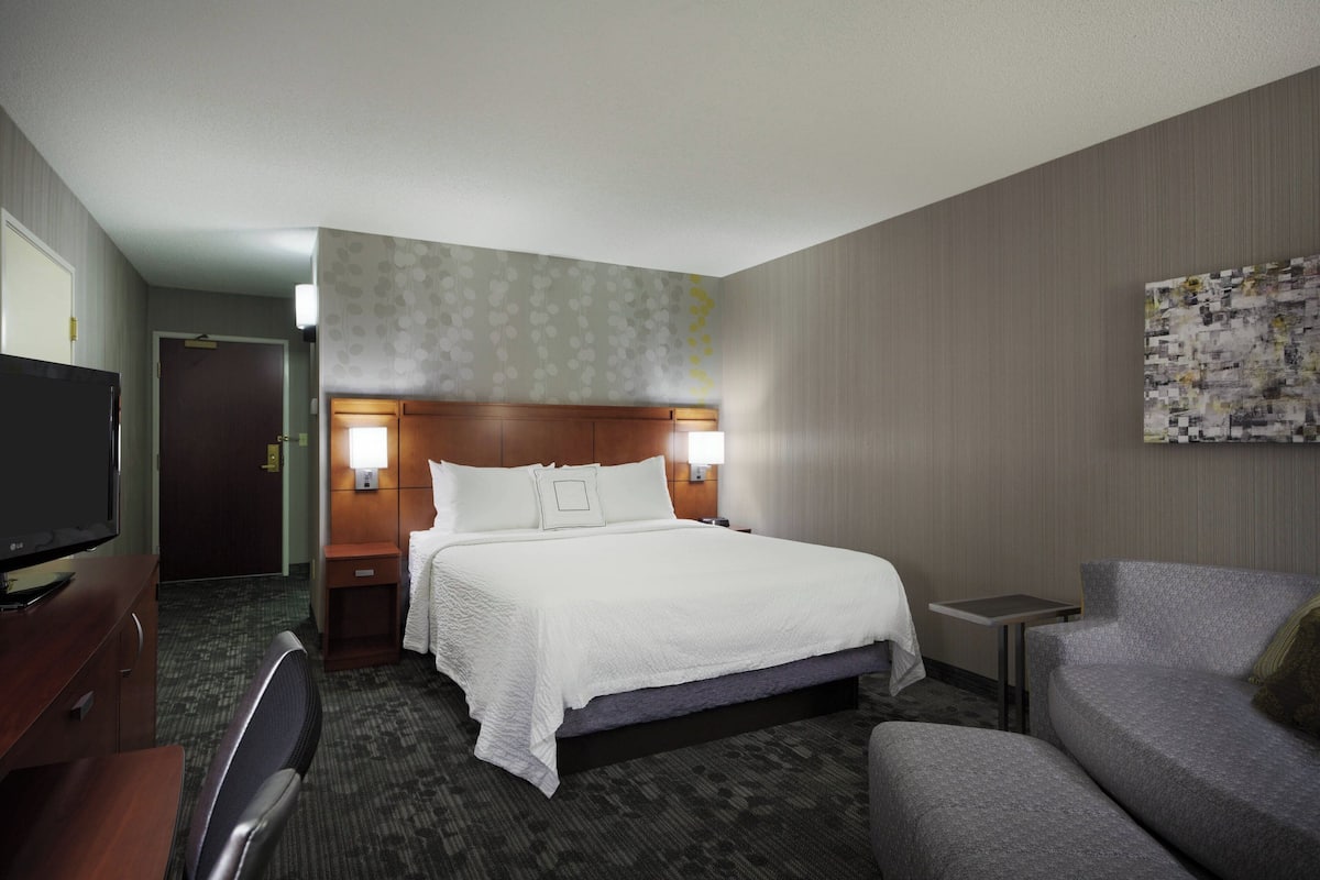 Quarto de hotel com cama, televisão, escrivaninha, cadeira e sofá.