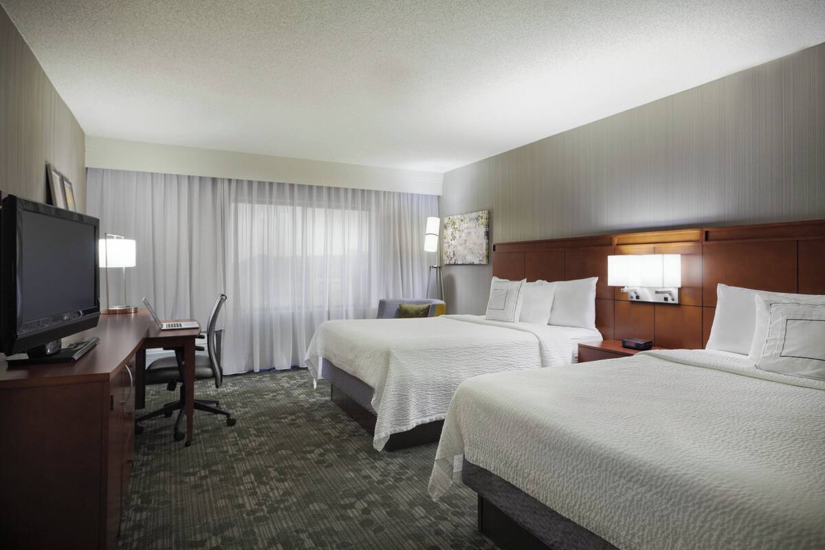 Quarto de hotel com duas camas, uma escrivaninha com um laptop, uma televisão e uma janela grande com cortinas.
