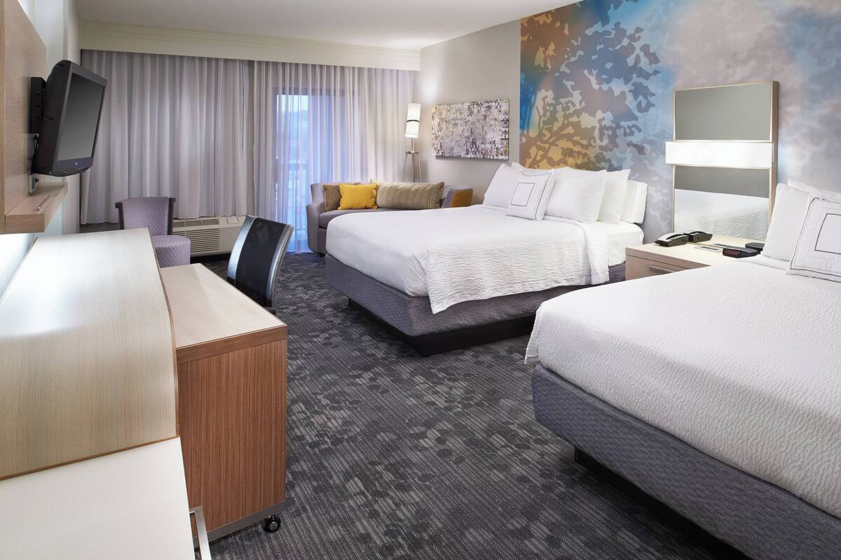 Una habitación de hotel con dos camas, un escritorio, una silla, un televisor y un ventanal con cortinas.