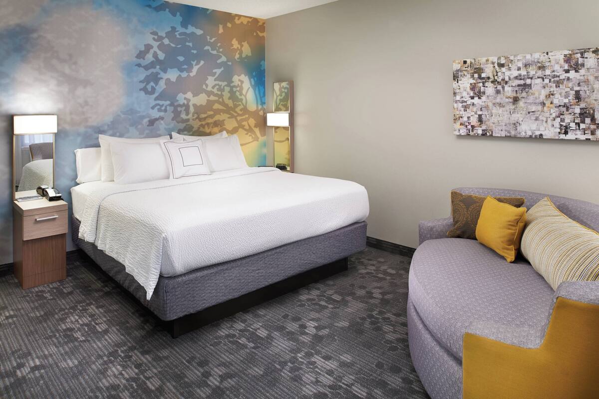 Habitación de hotel con una cama grande, un sofá, una mesita de noche y una pared decorada con arte abstracto.