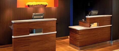 A reception area with two wooden reception desks, a digital screen, and a wall-mounted planter with yellow flowers.