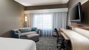 Habitación de hotel con cama, sofá, escritorio, silla, televisor y ventana con cortinas.