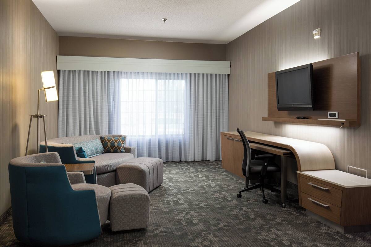 A hotel room with a sofa, ottoman, desk, chair, television, and a large window with curtains.