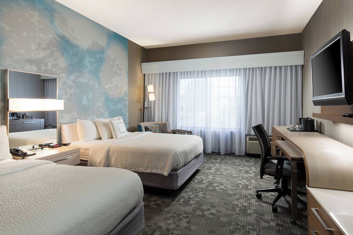 A hotel room with two beds, a desk, a chair, a TV, and a large window with curtains.