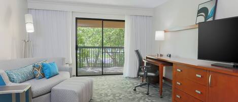 Ein Hotelzimmer mit grauem Sofa, einem kleinen Tisch, einem Schreibtisch mit Stuhl, einem Fernseher und einer Glasschiebetür, die auf einen Balkon führt.