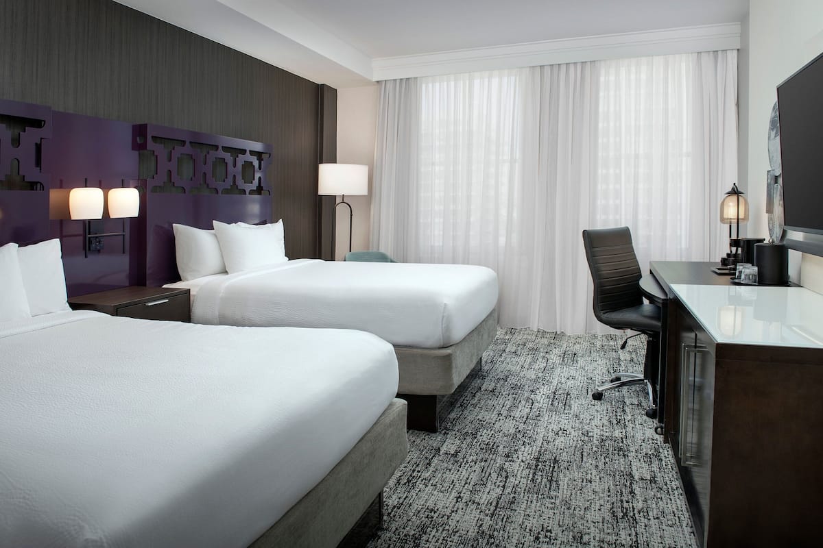 A hotel room with two beds, a desk, a chair, a lamp, and a large window with curtains.