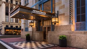 Entrada de un hotel con piso de cuadros, un letrero que dice "COURTYARD BY MARRIOTT" y dos plantas en macetas.