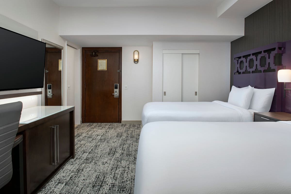 Une chambre d’hôtel comprenant un lit, un bureau avec un téléviseur à écran plat, une porte avec une serrure et un luminaire fixé au mur.