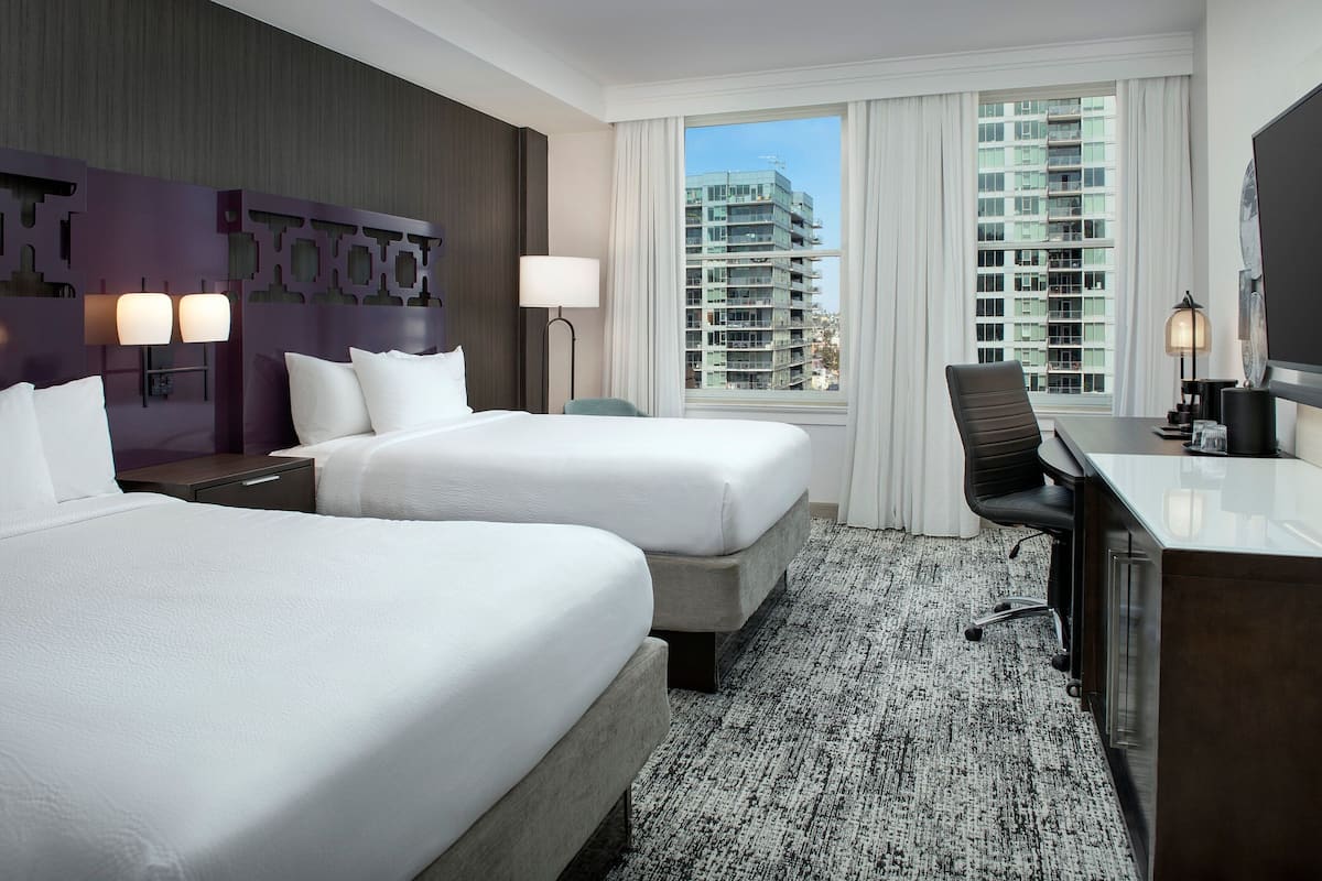 A hotel room with two beds, a desk, a chair, a large window with a view of buildings, and a patterned carpet.