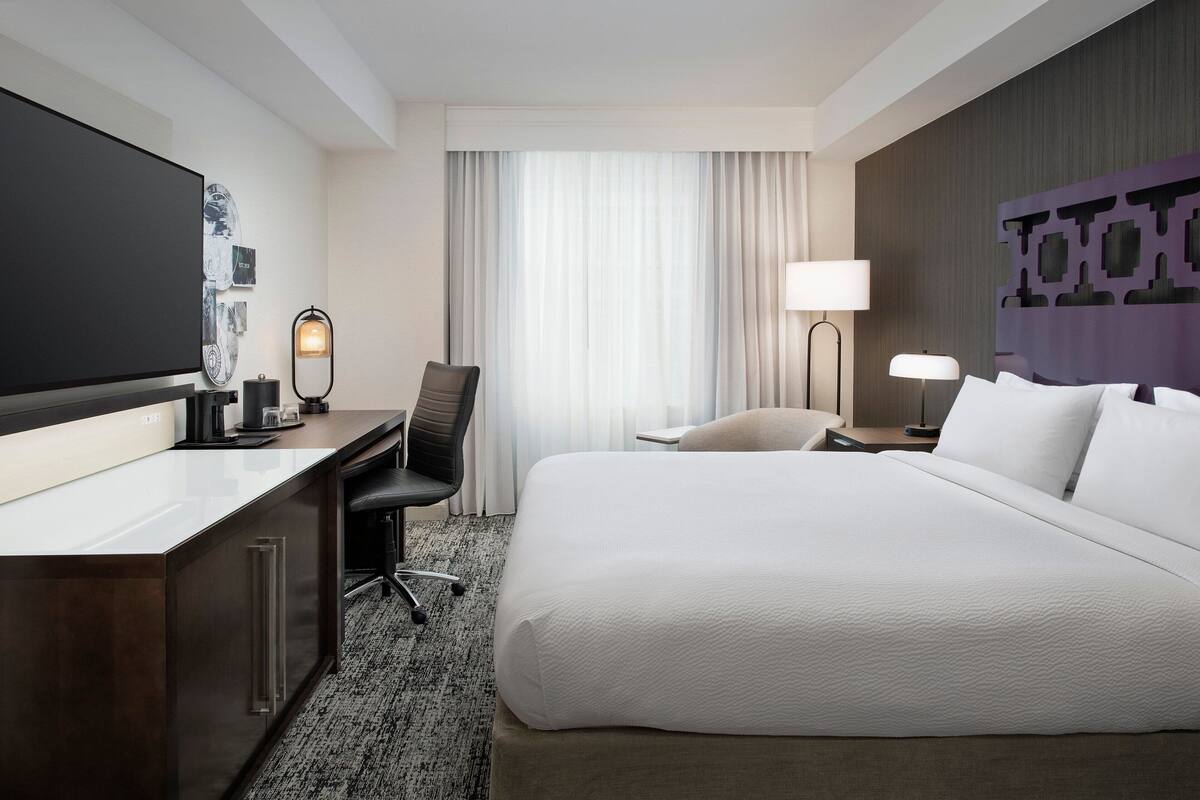 A hotel room with a large bed, a desk with a chair, a flat-screen TV, a lamp, and a window with curtains.