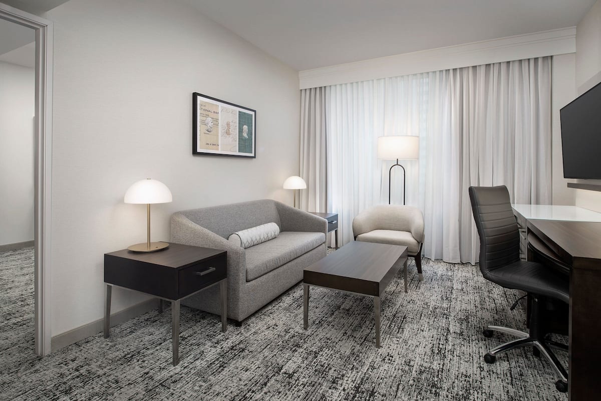 A hotel room with a grey sofa, a small table, a desk with a chair, two bedside lamps, a TV, and a framed picture on the wall.