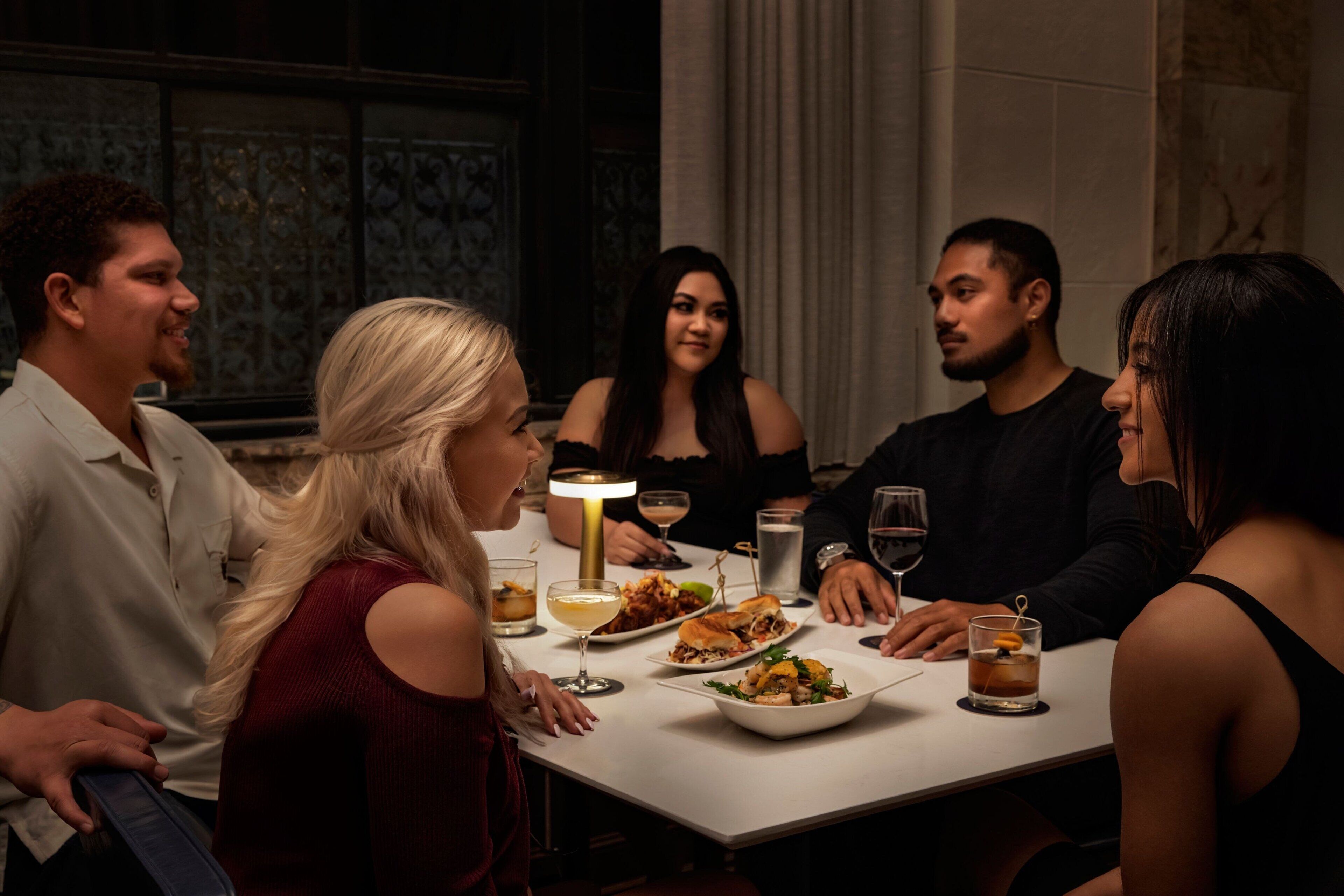 A group of people seated around a table with food and drinks, in a room with patterned walls and a chandelier.