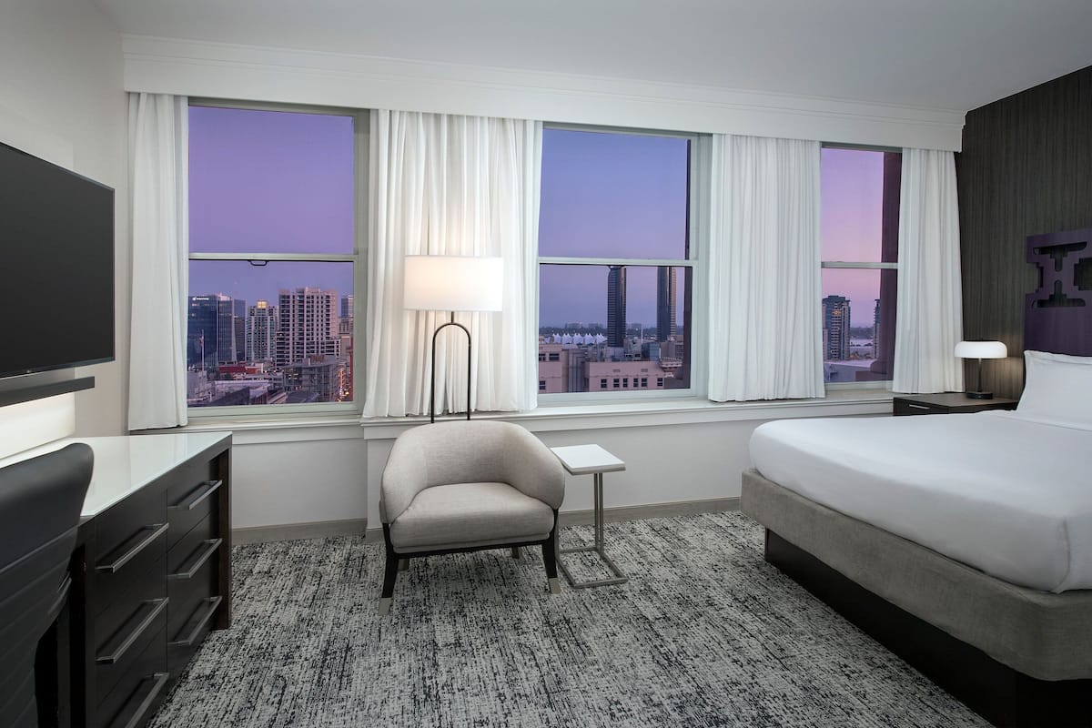 A hotel room with a bed, a desk, a chair, and a large window offering a city view.