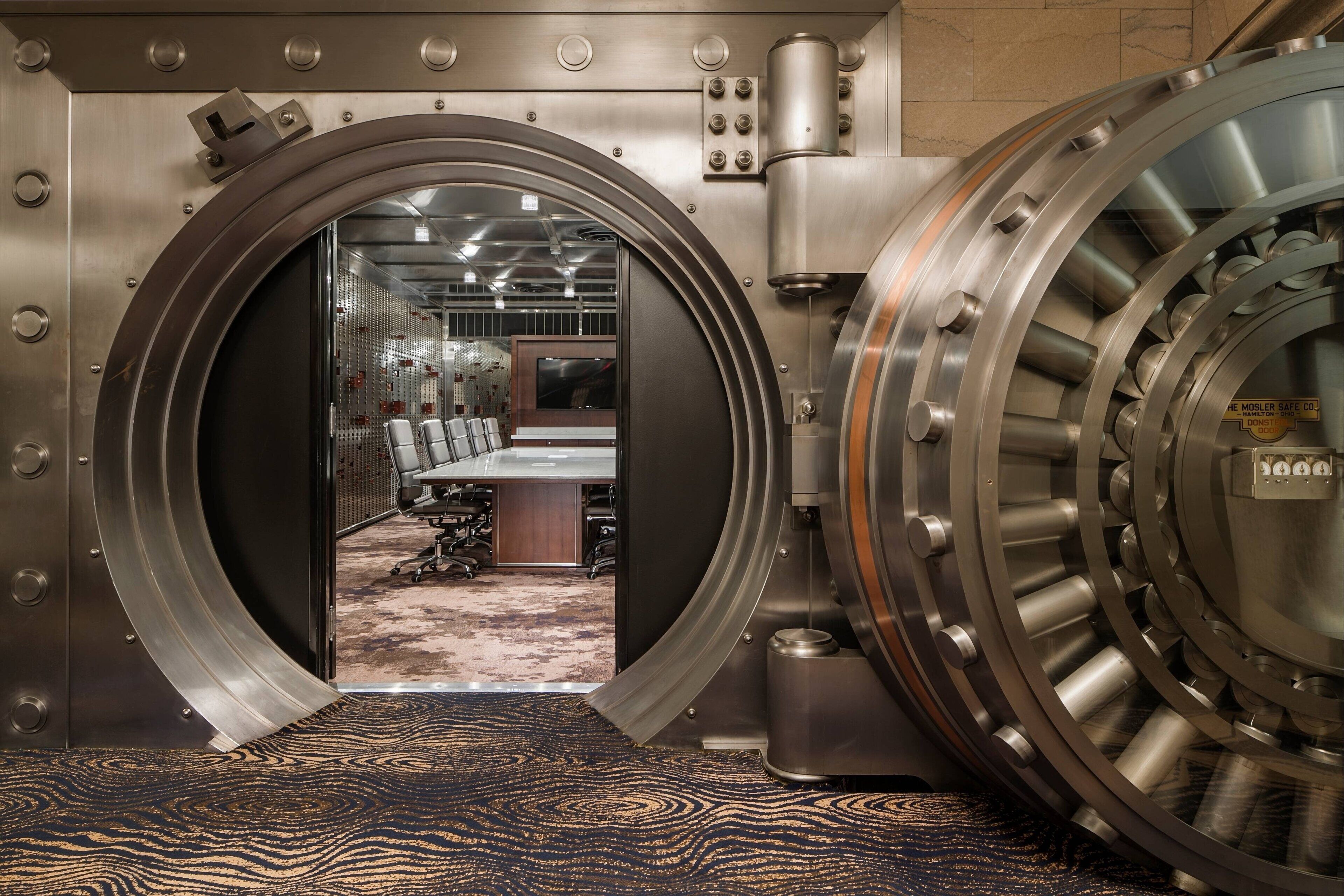 A large, metallic vault door with a view into a room containing a long table and chairs, suggesting a conference or meeting area.