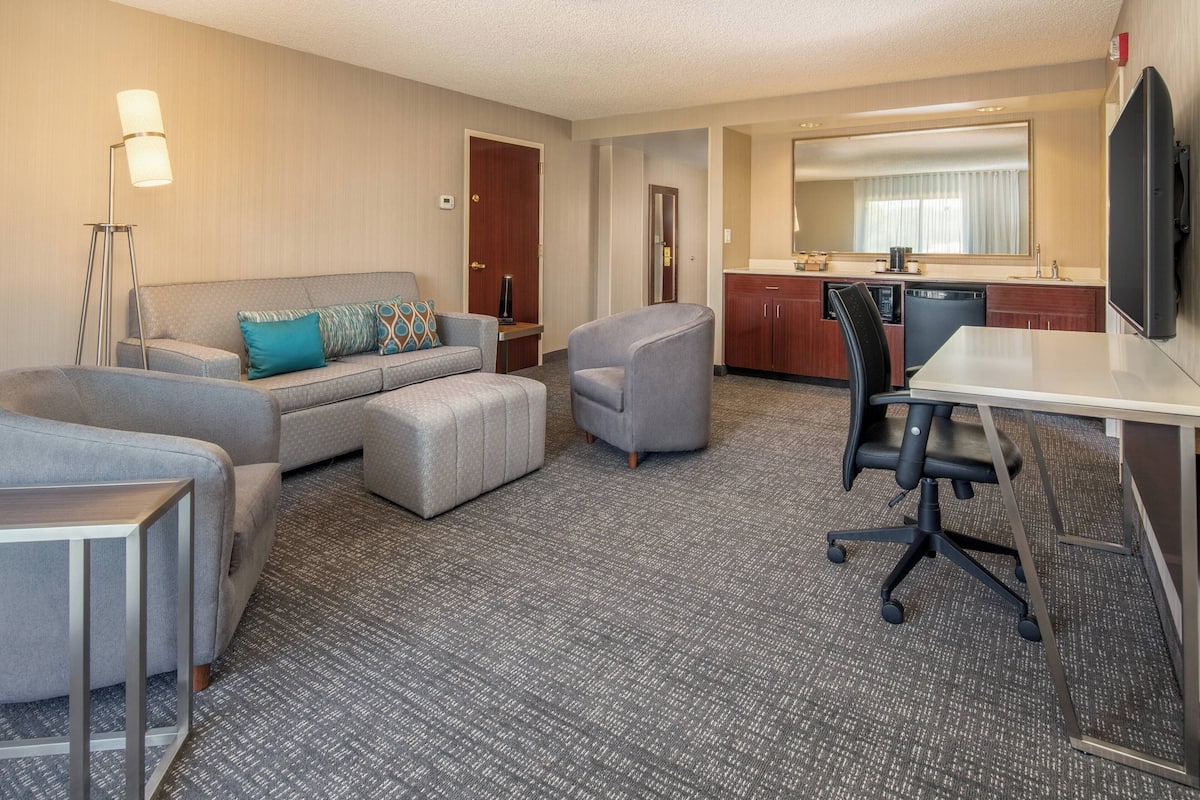 A hotel room with a grey sofa, a grey armchair, a small ottoman, a desk with a chair, a kitchenette, and a large mirror.