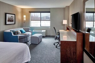 Habitación de hotel con cama, sofá, escritorio, silla, televisor y ventana con vistas a edificios.