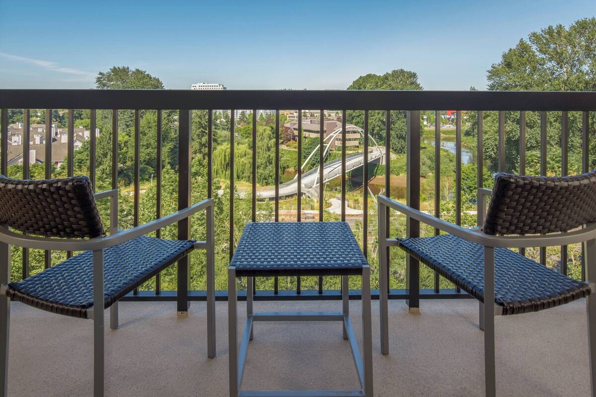 A balcony with two wicker chairs and a small table, overlooking a green landscape and a bridge.