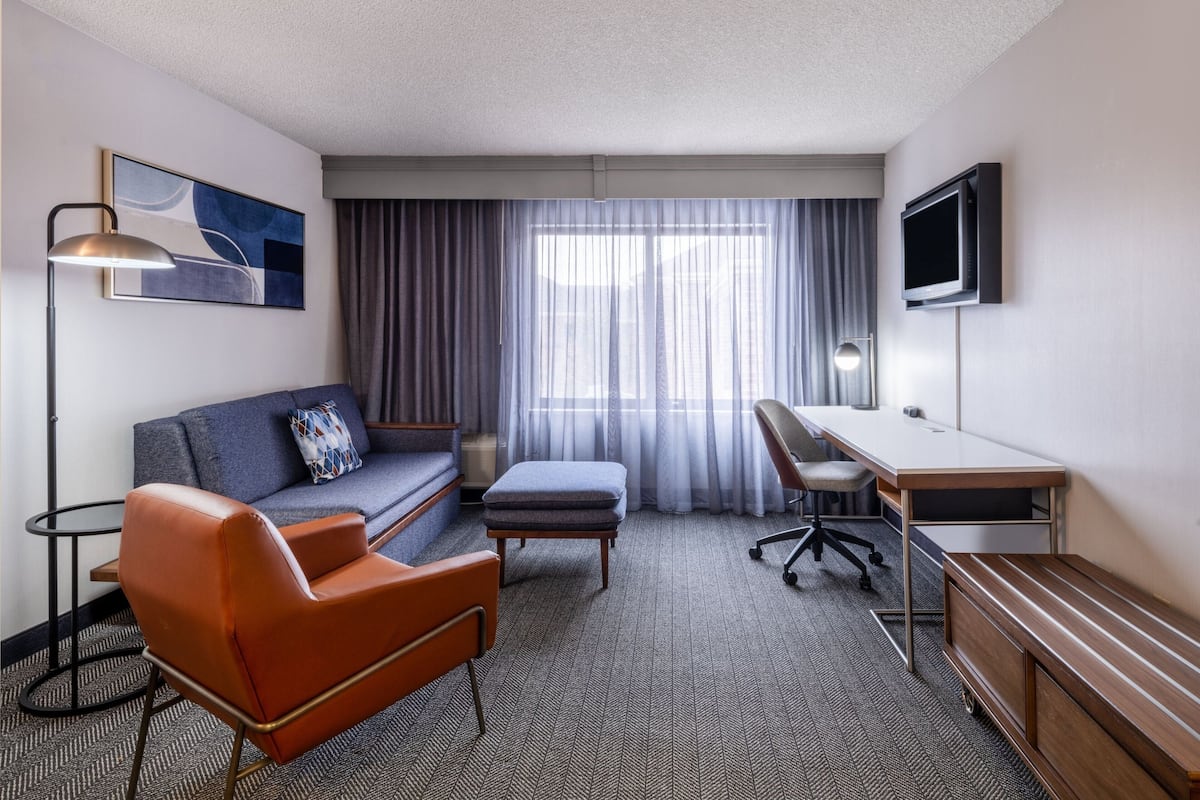 Ein Hotelzimmer mit Sofa, Ohrensessel, Hocker, Schreibtisch und Fernseher.