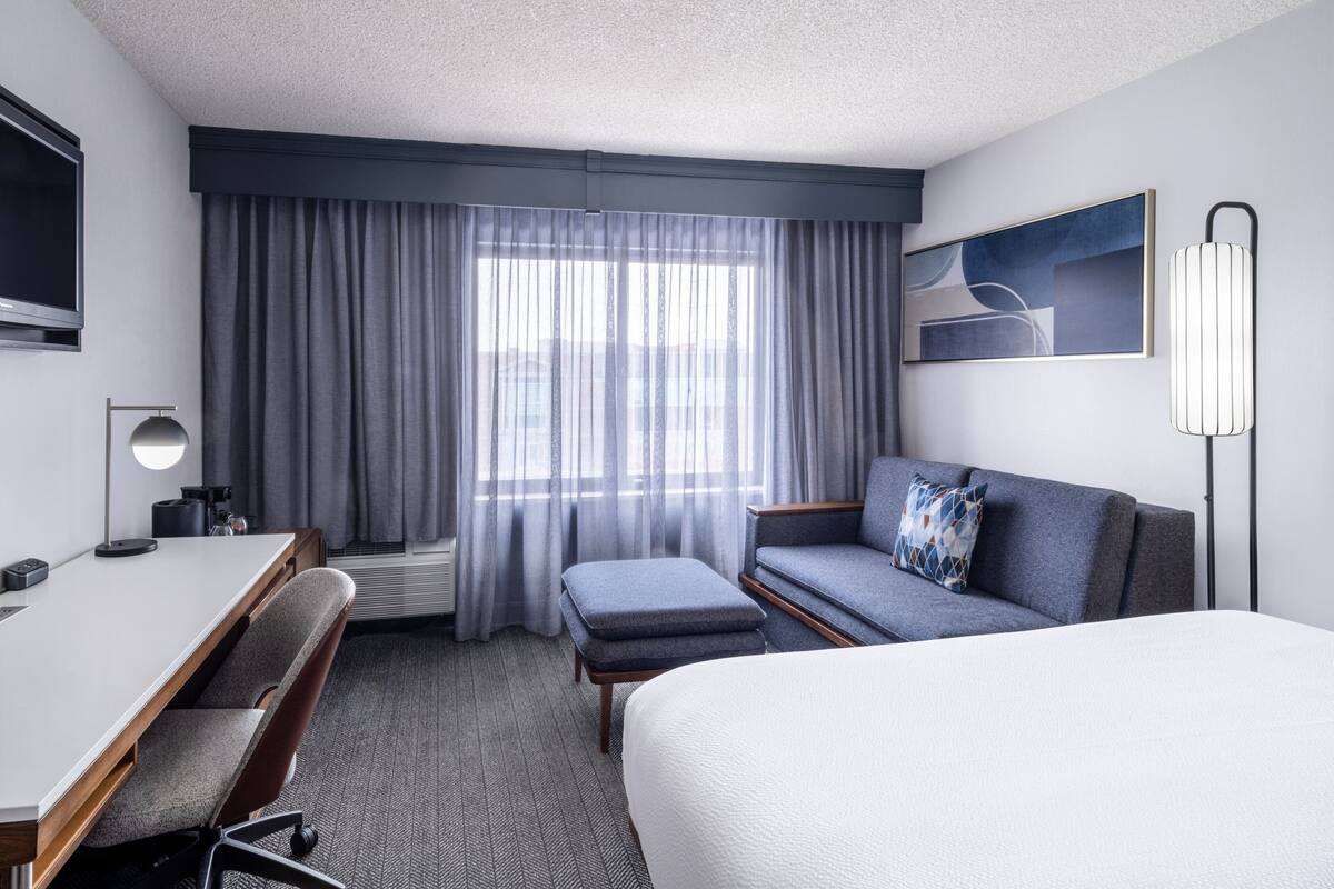 Ein Hotelzimmer mit Bett, Schreibtisch, Stuhl, Sofa, Fernseher, Lampe und einem Fenster mit Vorhängen.