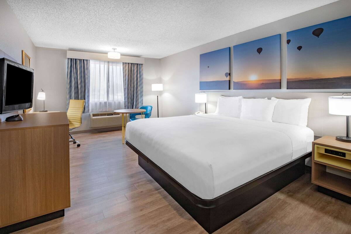 A hotel room with a large bed, a desk with a television, a chair, a desk lamp, and a wall with three paintings.
