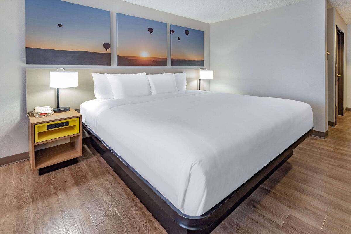 A neatly made bed with white linens, a bedside table with a lamp, and two wall-mounted paintings of hot air balloons at sunset.