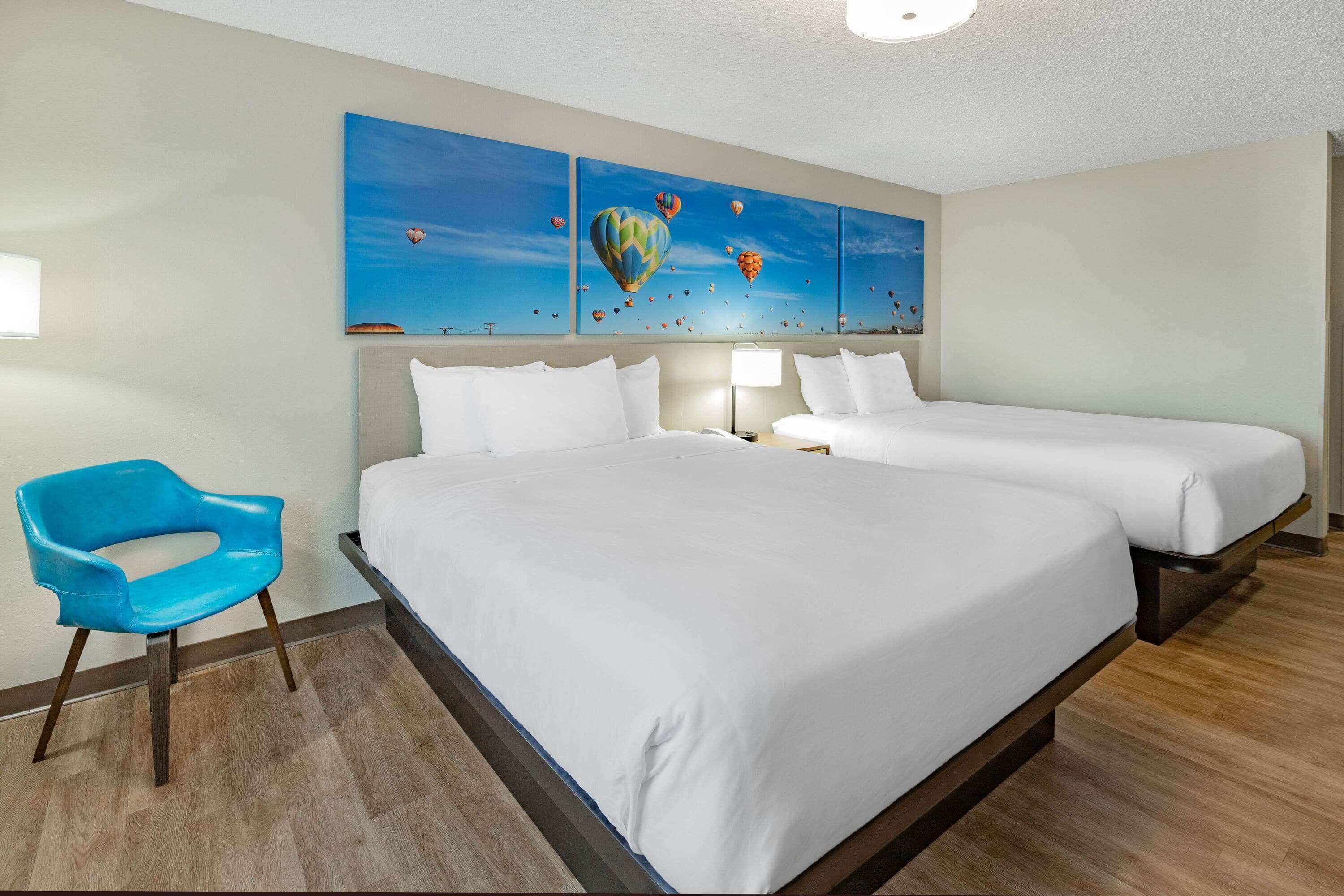 A hotel room with two beds, a blue chair, and a wall with three paintings of hot air balloons.