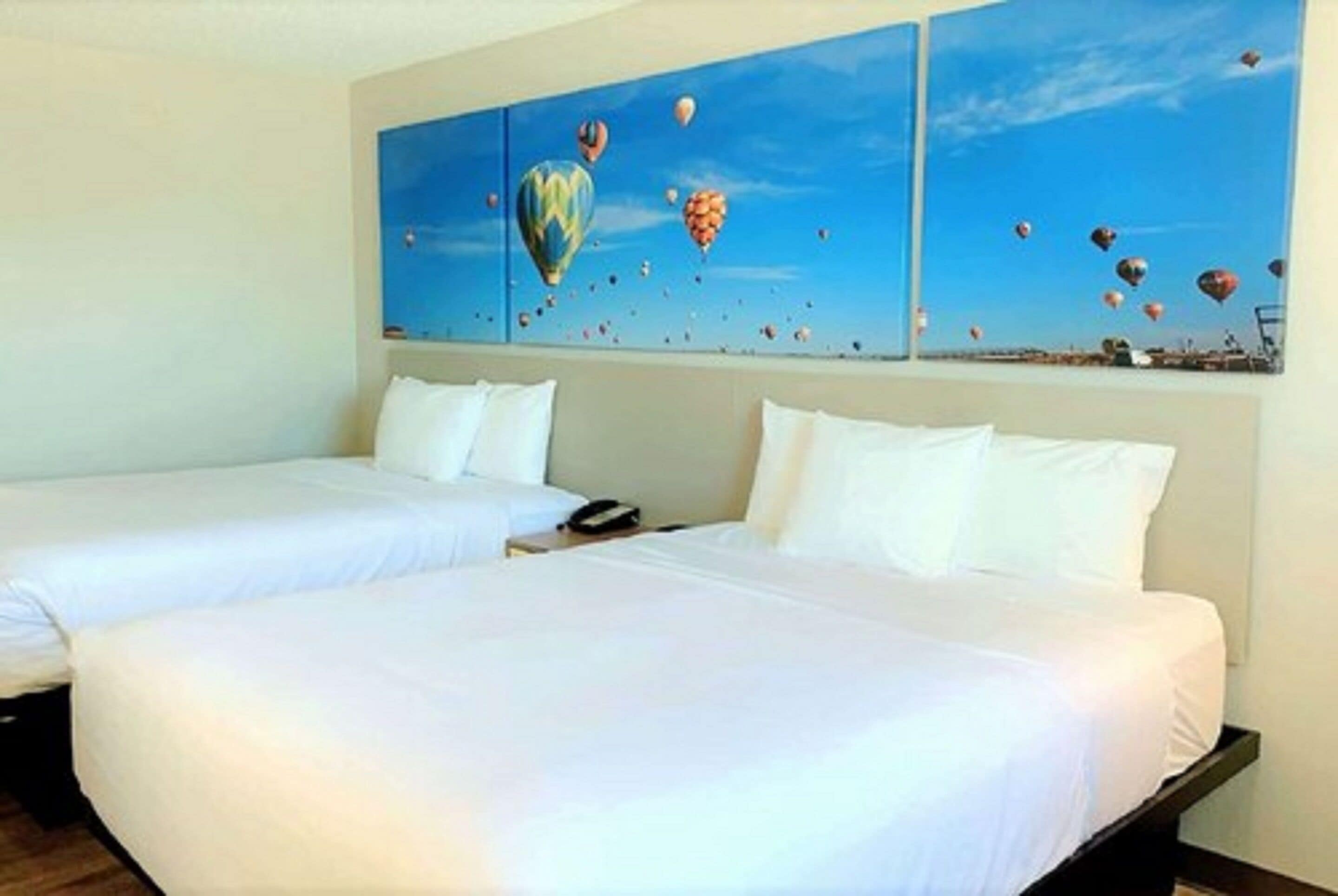 Habitación de hotel con dos camas, un cuadro grande de globos aerostáticos en la pared y una ventana con vistas al cielo.