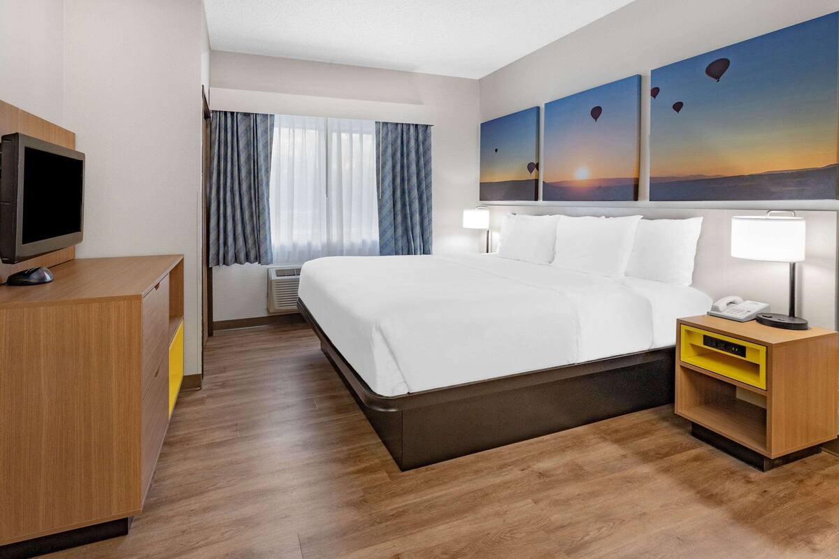 A hotel room with a large bed, a desk with a computer, a bedside table with a lamp, and a window with curtains.
