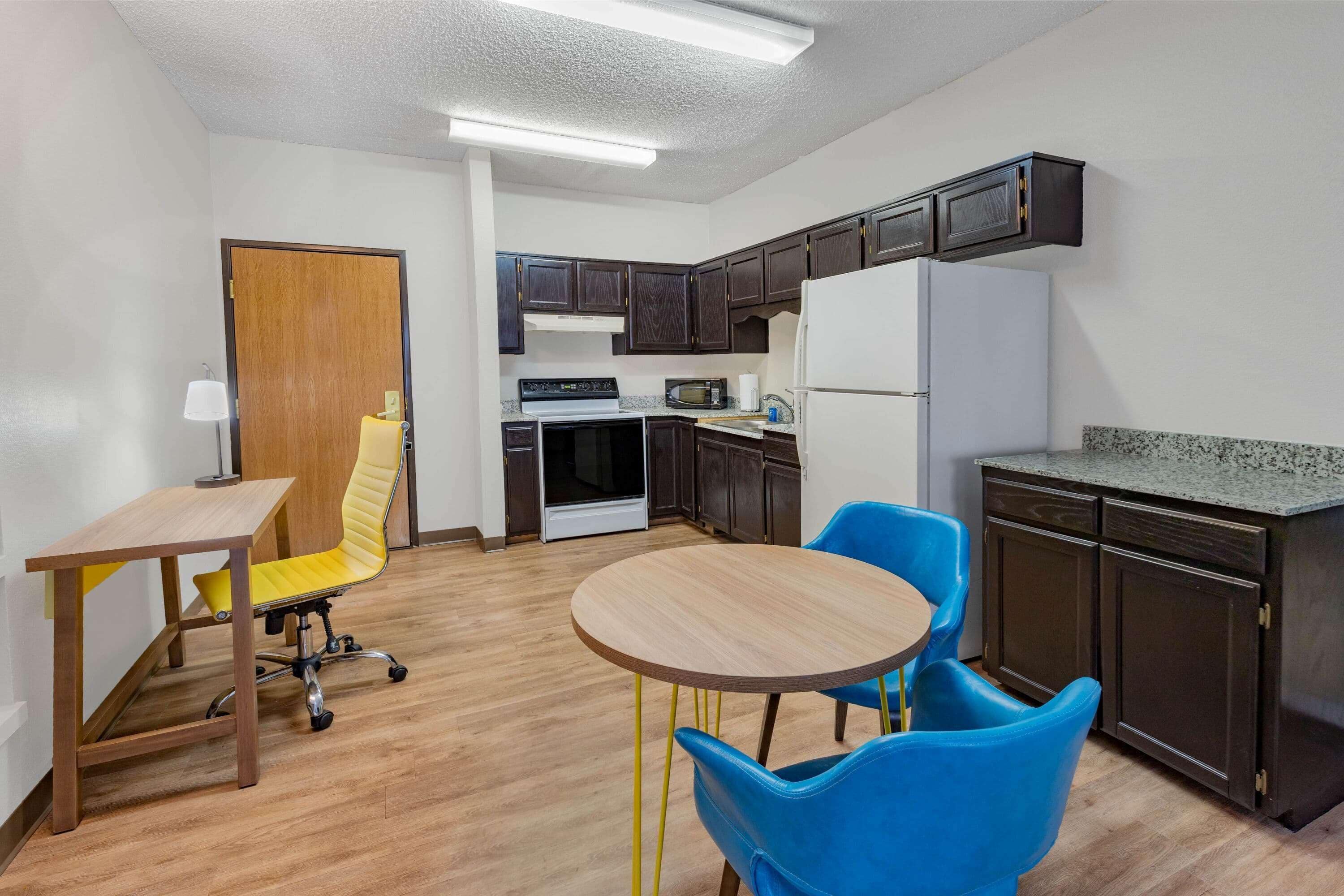 A compact kitchen with dark wooden cabinets, a white refrigerator, a microwave, and a stove. There is a small dining table with two chairs, one yellow and one blue.